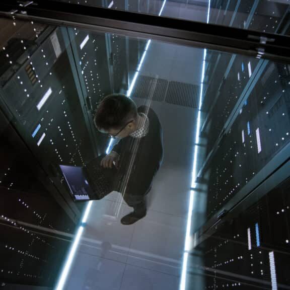 Top View Through the Glass of IT Engineer Working with Laptop in Data Center Full of  Active Rack Servers.